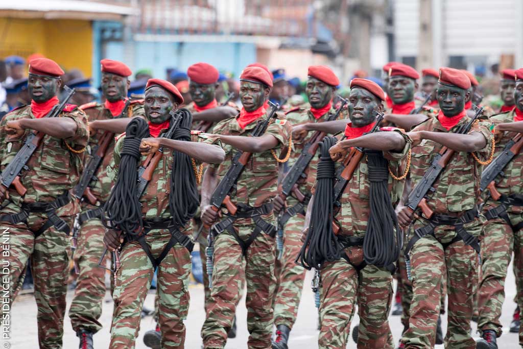 Un groupe de militaires en uniforme camouflage avec bérets rouges défile en formation dans une rue urbaine