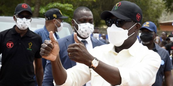 Un homme en chemise blanche et casquette noire fait un pouce levé entouré d'autres hommes portant des masques de protection
