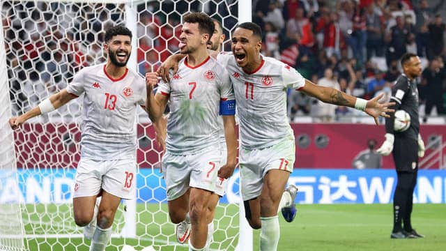 Trois joueurs en maillots blancs célèbrent un but devant le but avec le gardien adverse en arrière-plan.