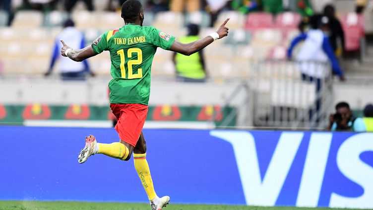 Un joueur en maillot vert, short rouge et chaussettes jaunes célèbre un but avec les bras écartés sur un terrain de football