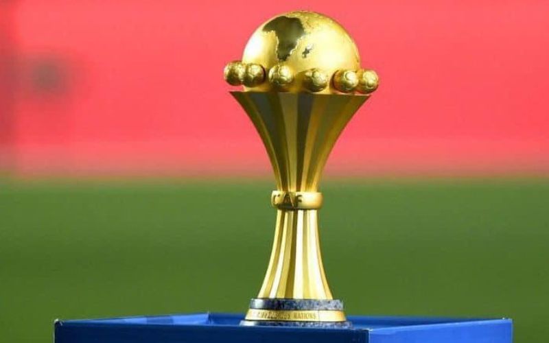 Trophée doré de la Coupe d'Afrique des Nations posé sur un socle bleu devant un fond rouge flou