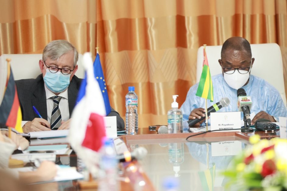 Deux hommes masqués assis à une table avec des drapeaux, des bouteilles d'eau et un flacon de gel hydroalcoolique