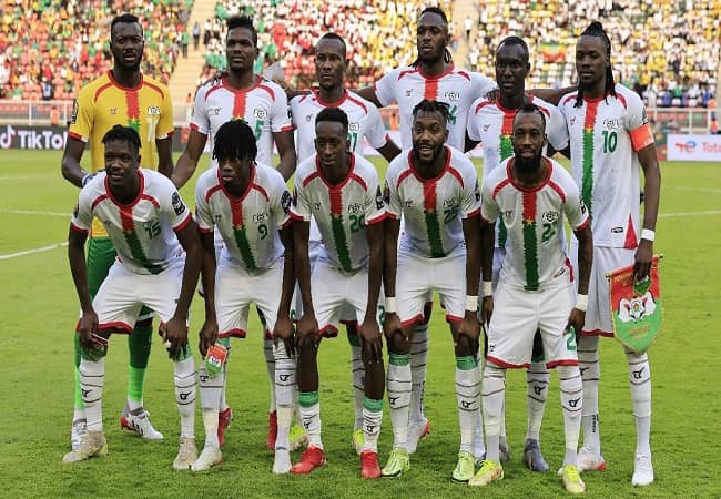 Onze joueurs de football en tenue blanche avec bandes rouge et verte posent devant un stade rempli de spectateurs