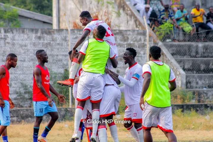 Joueurs en maillots blanc et rouge célébrant un but tandis que des joueurs en maillots rouges et bleus s'éloignent sur le terrain de football
