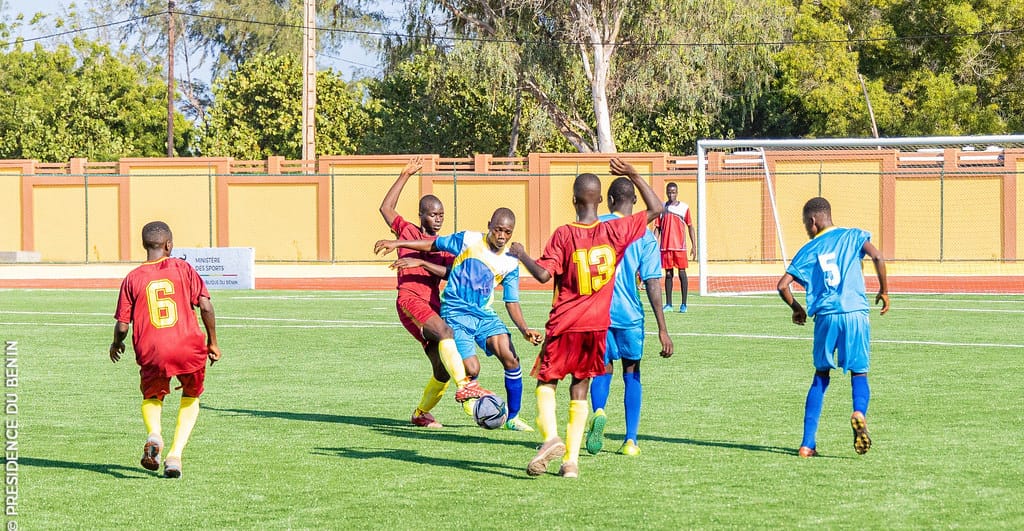 Jeunes joueurs en maillots rouges et bleus disputant un ballon sur un terrain de football en gazon synthétique devant un mur ocre