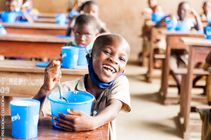 Un enfant souriant mange dans une cantine scolaire avec d’autres enfants assis à des pupitres en arrière-plan