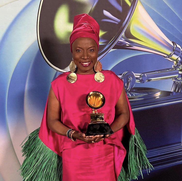 Femme souriante en robe rose fuchsia et grand foulard assorti tenant un trophée devant un grand décor de gramophone doré