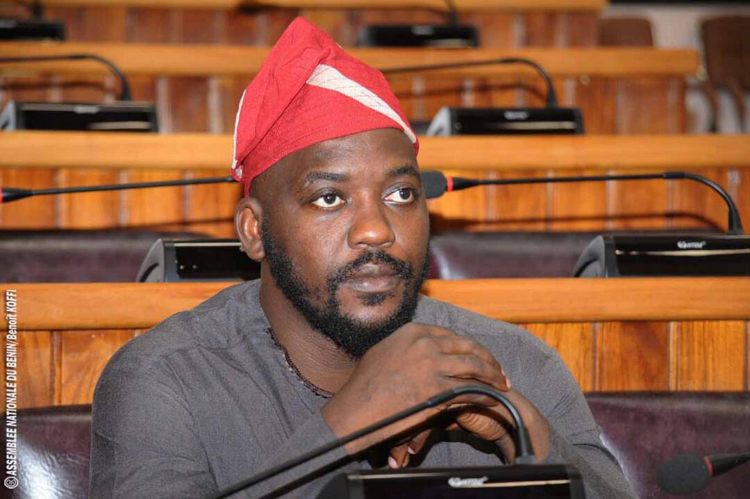 Un homme barbu portant un bonnet rouge et un vêtement gris assis dans une salle avec des bancs en bois et des micros devant lui