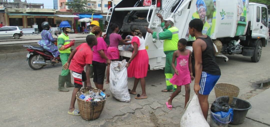 Des adultes et enfants déposent des sacs de déchets devant un camion de ramassage en tenue de travail verte sur une route urbaine