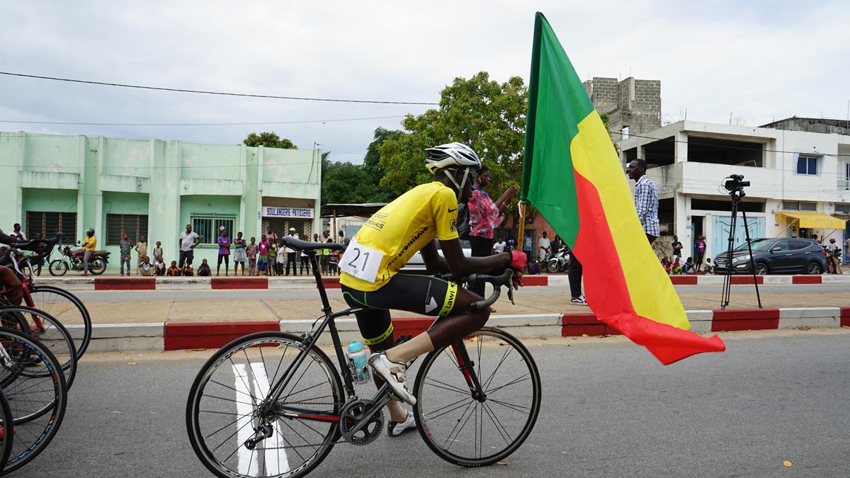 Un cycliste en tenue jaune et casque porte un drapeau tricolore rouge, jaune et vert lors d'une course en ville