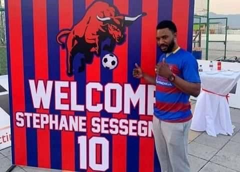 Un homme en tenue de sport fait un pouce levé devant une affiche aux couleurs rouges et bleues avec un taureau et un message de bienvenue