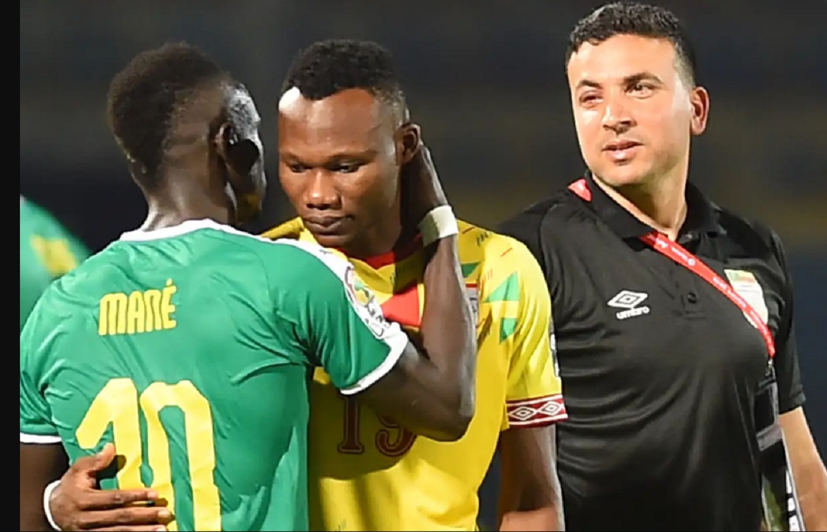 Deux joueurs de football, l’un en maillot vert numéro 10, l’autre en maillot jaune numéro 19, s’embrassent sur la joue avec un arbitre à leurs côtés