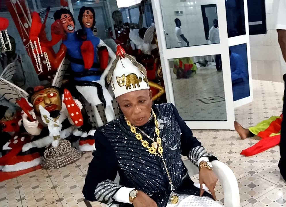 Homme âgé en tenue traditionnelle avec colliers et coiffe assis devant des masques et sculptures colorés dans une pièce intérieure
