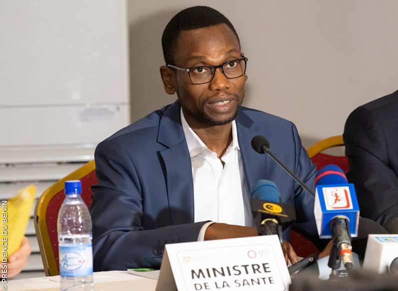 Homme en costume bleu parlant au micro lors d une conférence avec panneaux et bouteilles sur la table