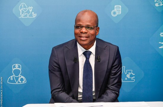 Un homme en costume sombre et cravate sourit assis devant un fond bleu orné d’icônes blanches liées à la santé et l’économie