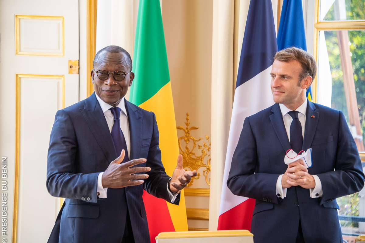 Deux hommes en costume discutent devant deux drapeaux dont un tricolore français dans une salle élégante avec une grande fenêtre
