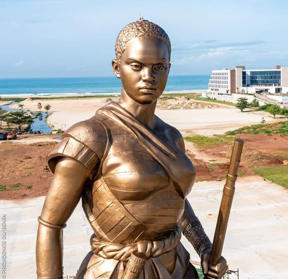 Statue monumentale en bronze d’une femme armée dans un paysage côtier avec bâtiments en arrière-plan
