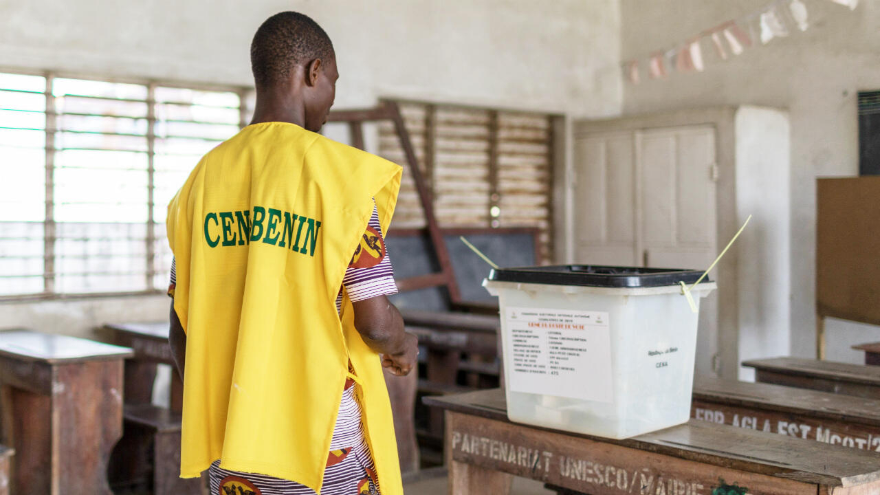 Un homme portant un gilet jaune marqué est en train de déposer un bulletin dans un urne transparente dans une salle de vote