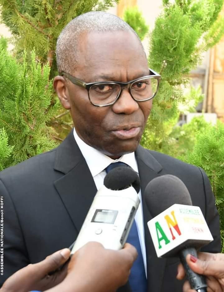 Homme en costume sombre et lunettes répond à plusieurs micros tenus devant lui avec un fond de verdure dense