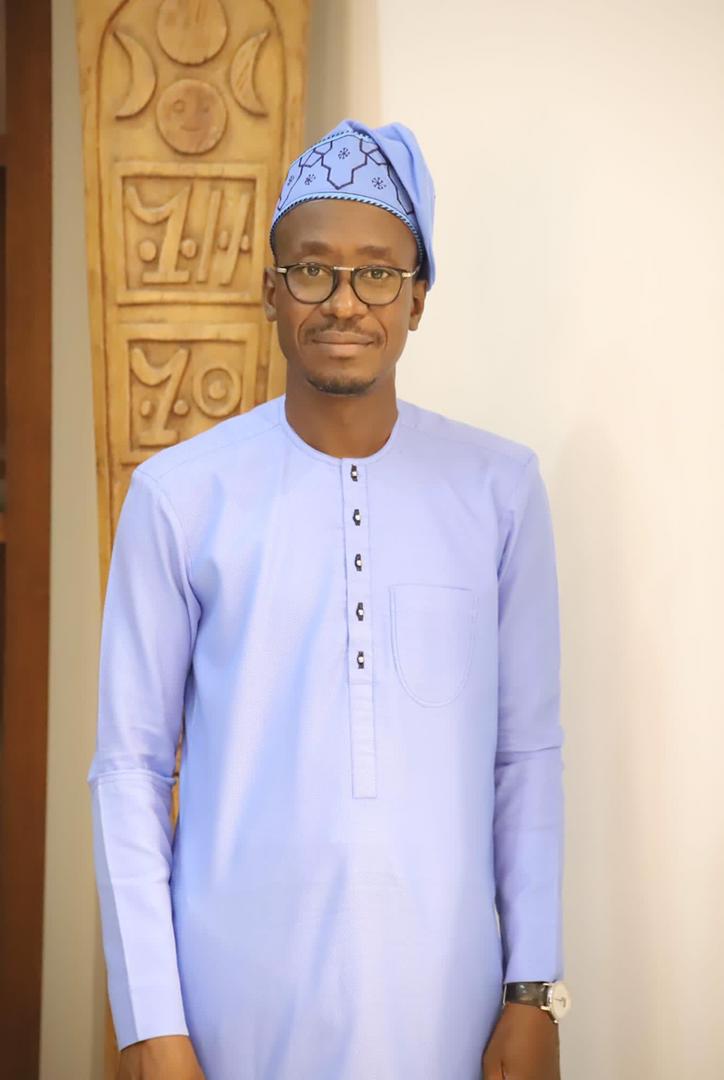 Homme souriant portant une tenue traditionnelle bleue et un bonnet assorti devant un mur clair et un panneau sculpté en bois