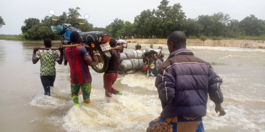 Quatre personnes portant des sacs et une moto traversent un cours d’eau en crue entouré d’arbres et de végétation