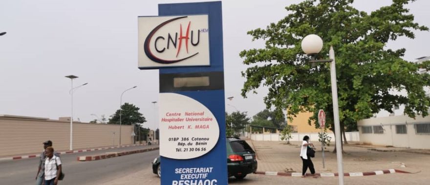 Panneau d'entrée du Centre National Hospitalier Universitaire avec quelques personnes et voitures autour