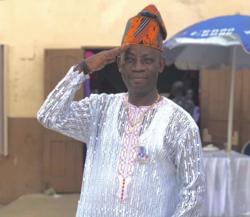 Un homme souriant en tenue traditionnelle blanche et chapeau orange fait un salut militaire devant un bâtiment beige.