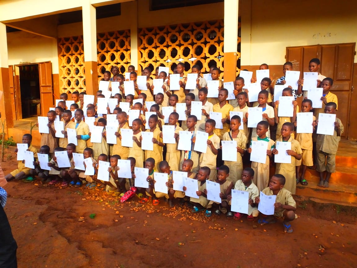 Groupe d’enfants en uniforme beige posant en rangées avec des documents blancs devant un bâtiment ocre aux murs ajourés