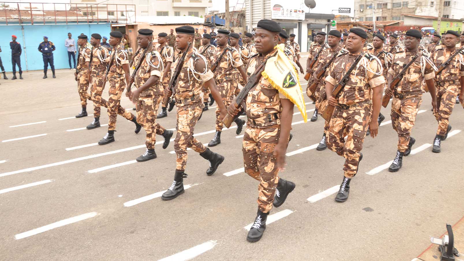 Des militaires en uniforme camouflé défilent en formation serrée sur une voie urbaine lors d'une cérémonie extérieure