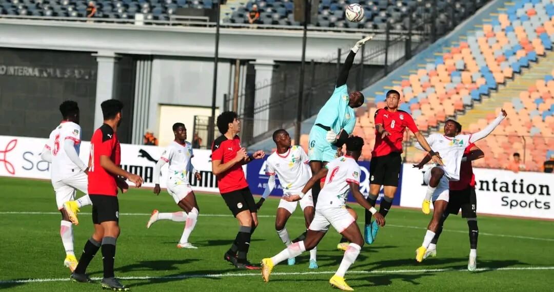 Un joueur en combinaison bleue saute pour attraper un ballon devant plusieurs joueurs en tenues rouges et blanches sur un terrain de football.