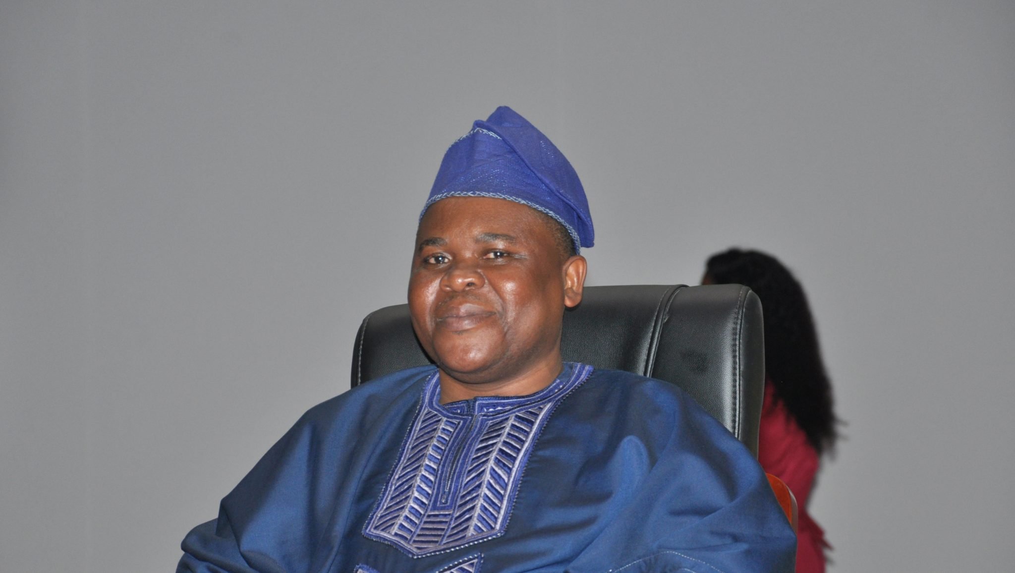 Homme souriant en tenue traditionnelle bleue assis sur une chaise devant un fond gris