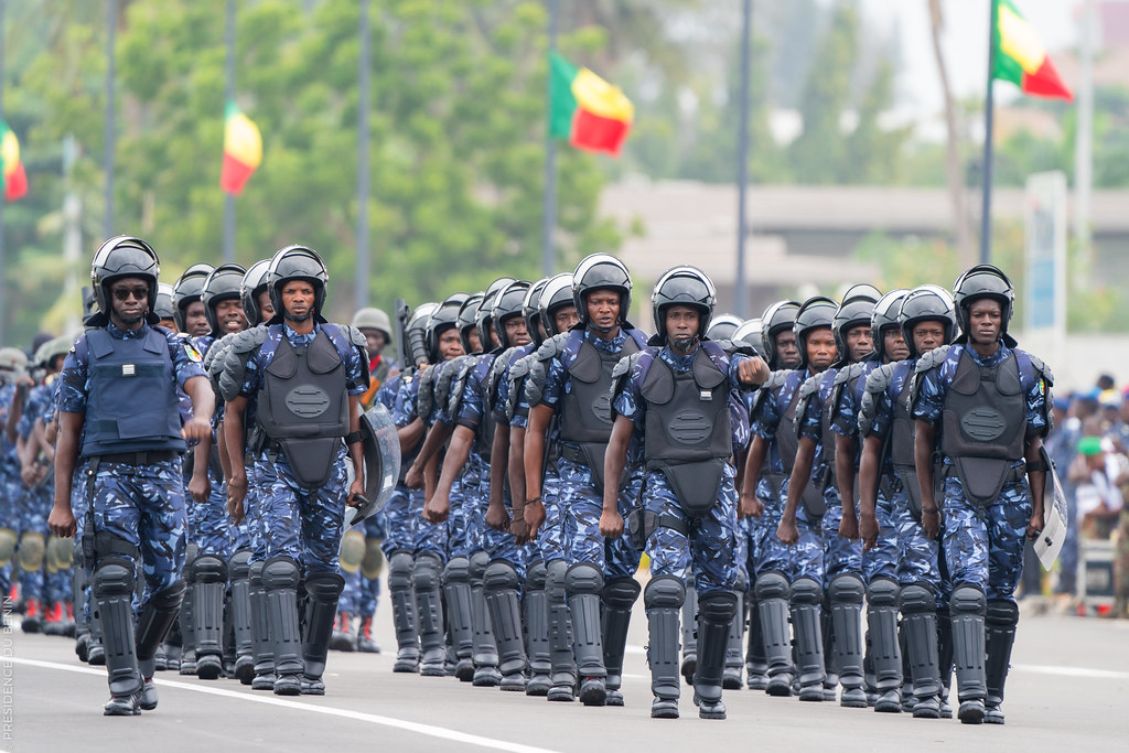 Des agents en tenue tactique et casque défilent en formation serrée sur une avenue bordée de drapeaux colorés