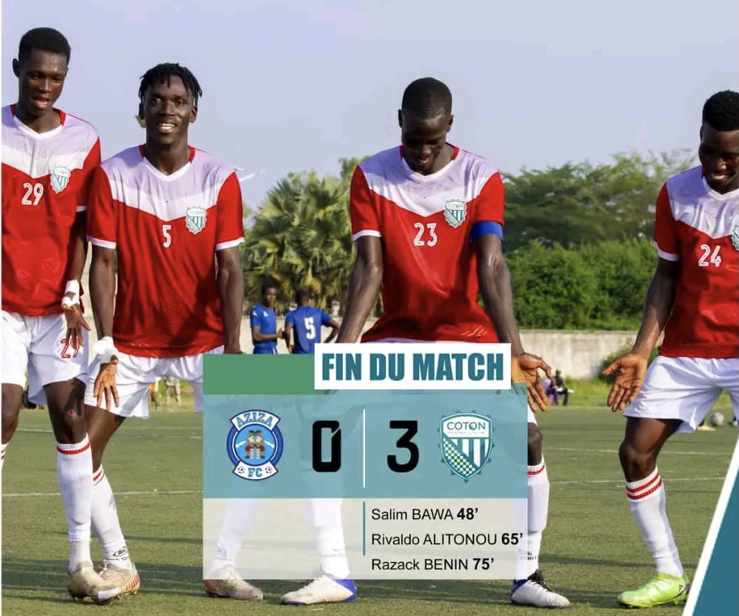 Quatre joueurs en maillots rouges et blancs posent souriants sur un terrain de football gazonné sous un ciel clair