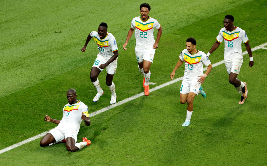 Quatre joueurs en maillot blanc avec bandes vert, jaune et rouge célèbrent un but sur un terrain de football vert