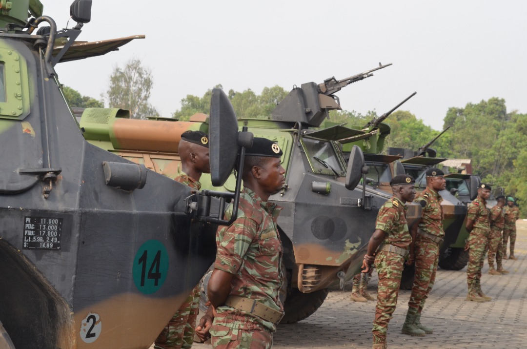 Des militaires en tenue de camouflage se tiennent debout en ligne devant plusieurs véhicules blindés stationnés sur un sol pavé
