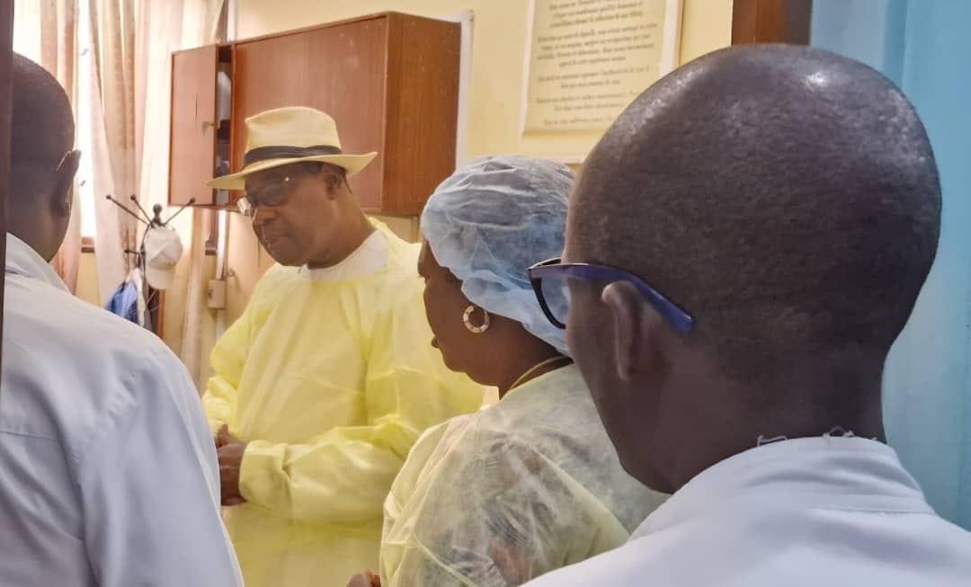 Quatre personnes en tenue médicale discutent dans une pièce, l’une portant un chapeau et une blouse jaune
