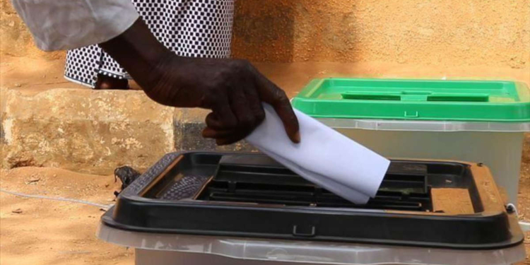Une main dépose un bulletin dans une urne pour le vote devant un mur ocre et une autre urne verte en arrière-plan