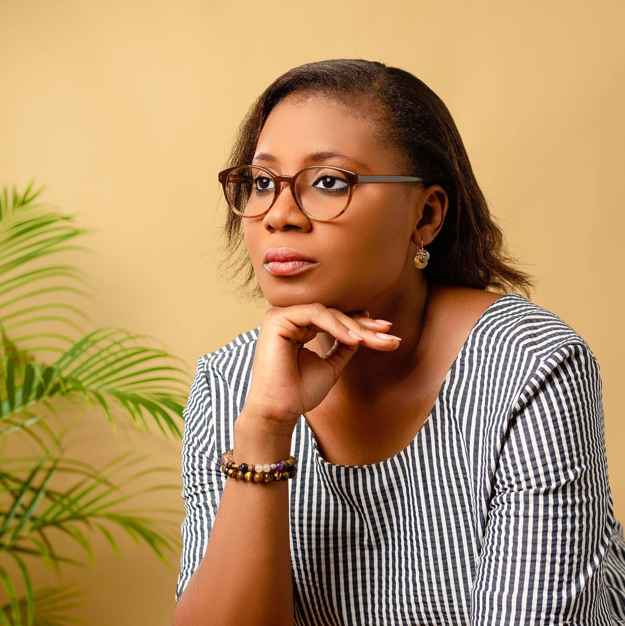 Femme portant des lunettes posant son menton sur la main avec un fond beige et une plante verte sur la gauche