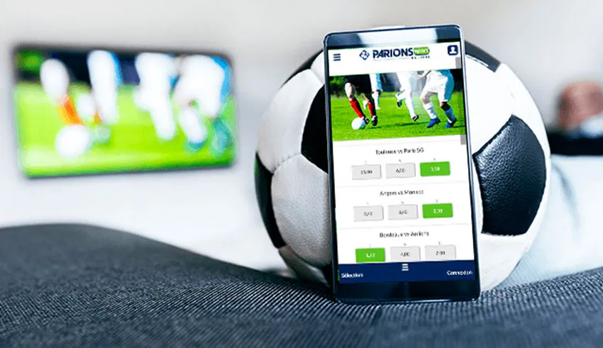 Téléphone affichant une application de paris sportifs appuyé sur un ballon de football avec un écran flou en arrière-plan montrant un match