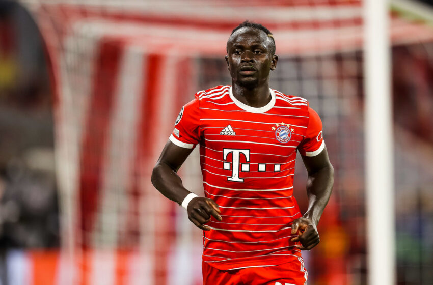 Joueur de football en maillot rouge avec sponsors, sur un terrain devant un but flou en arrière-plan