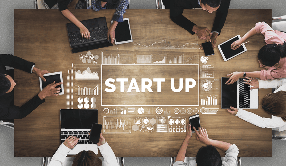 Plusieurs personnes autour d'une table en bois utilisent ordinateurs portables, tablettes et smartphones avec affichage digital START UP au centre