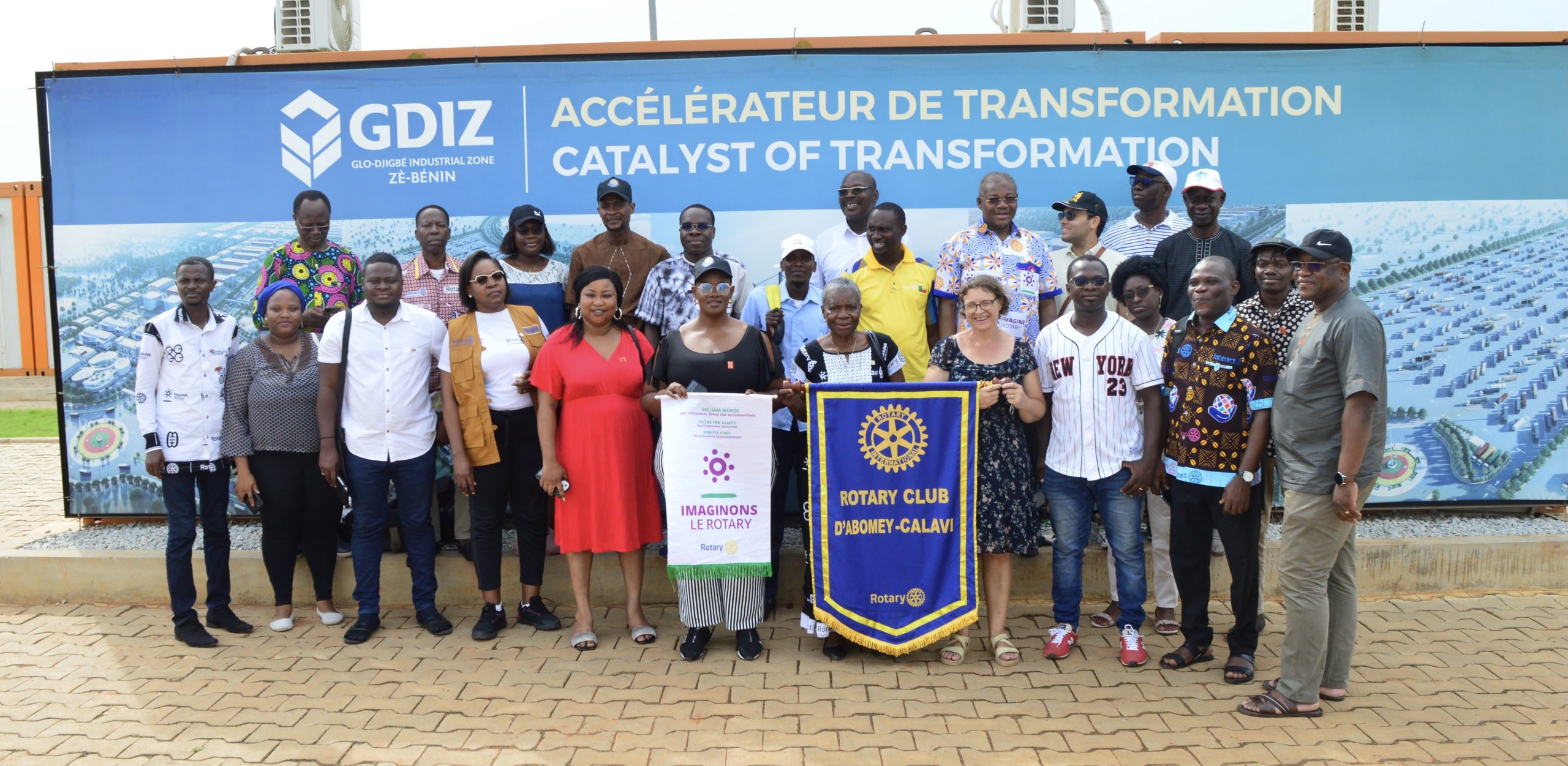 Groupe de personnes posant devant un panneau bleu avec des banderoles de deux clubs de Rotary Clubs et des inscriptions en anglais et français