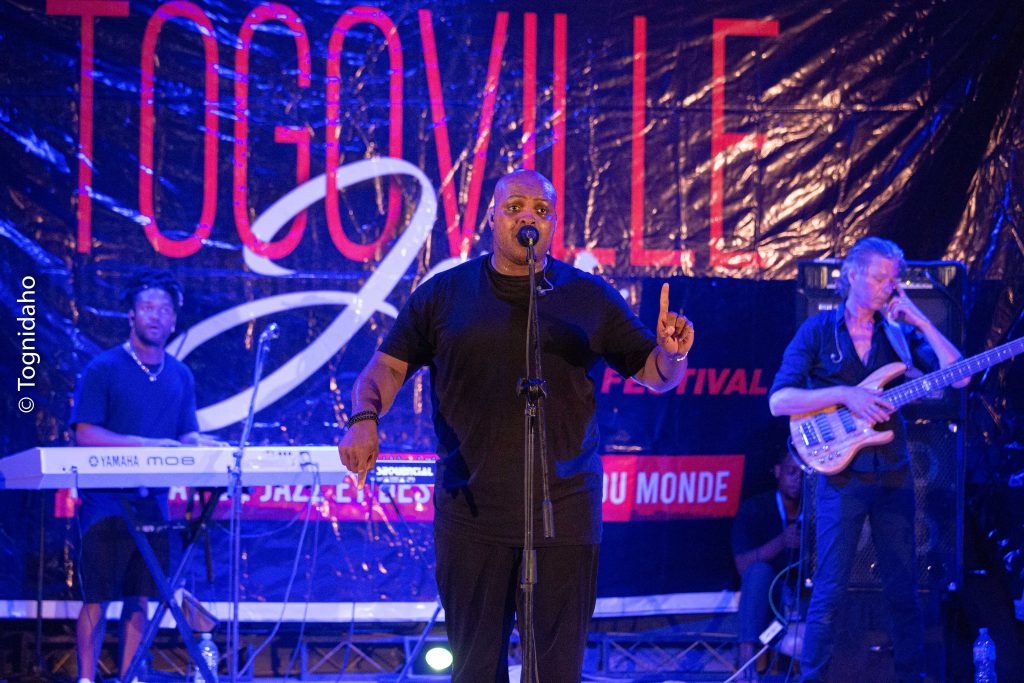 Un chanteur au centre sur scène entouré de musiciens, devant une grande bannière du festival Togoville Jazz.
