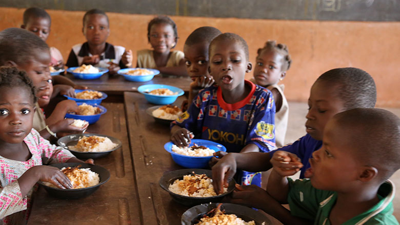 Des enfants assis à une longue table en bois mangent dans des assiettes de nourriture dans une salle de classe