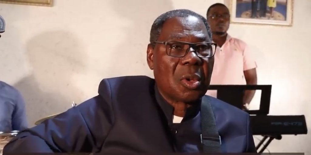 Homme portant des lunettes et une veste sombre chante avec un groupe en arrière-plan dans une pièce éclairée
