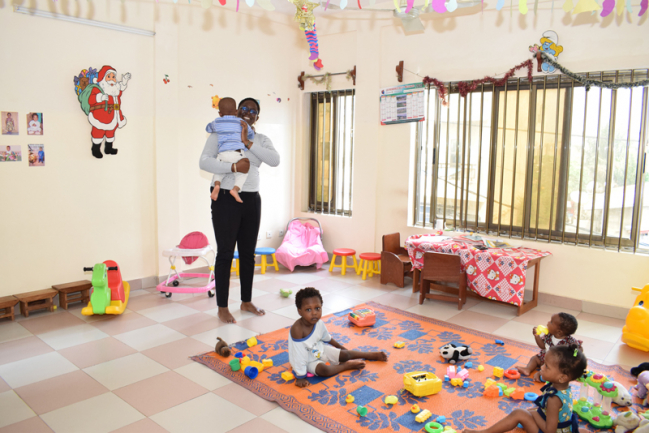 Une femme portant un enfant dans une salle de jeux avec plusieurs tout-petits assis sur un tapis parmi des jouets colorés