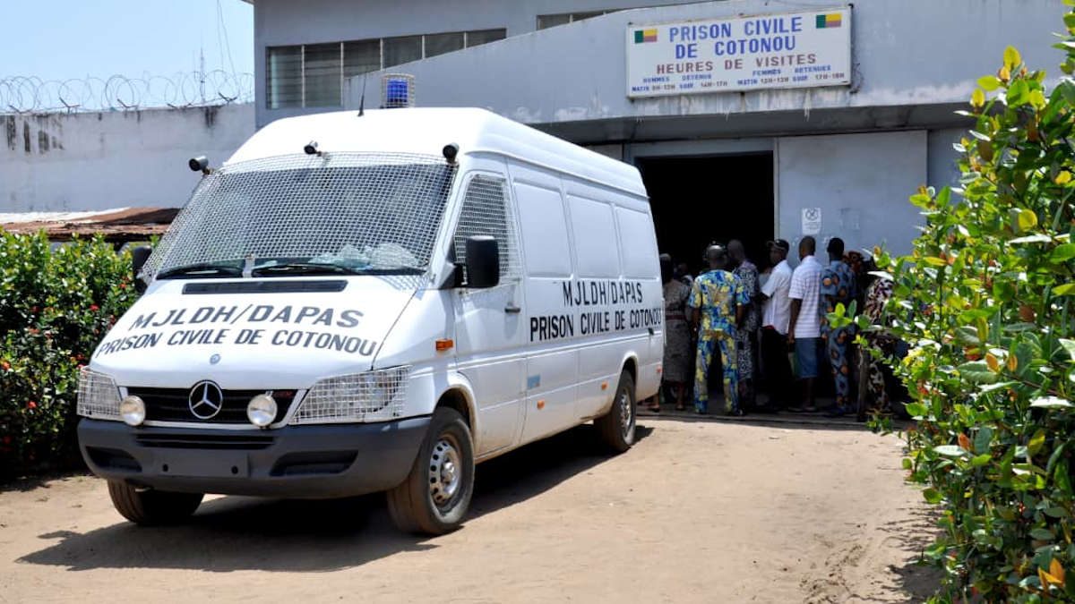 Fourgon blanc avec grillage aux fenêtres stationné devant l'entrée d'une prison entourée de murs barbelés avec un groupe de personnes à proximité