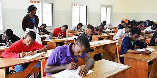 Des élèves concentrés écrivent un examen dans une salle de classe lumineuse avec un surveillant debout au fond