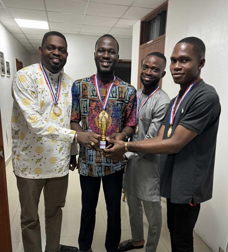 Quatre hommes souriants portent des médailles et tiennent un trophée doré ensemble dans un couloir intérieur