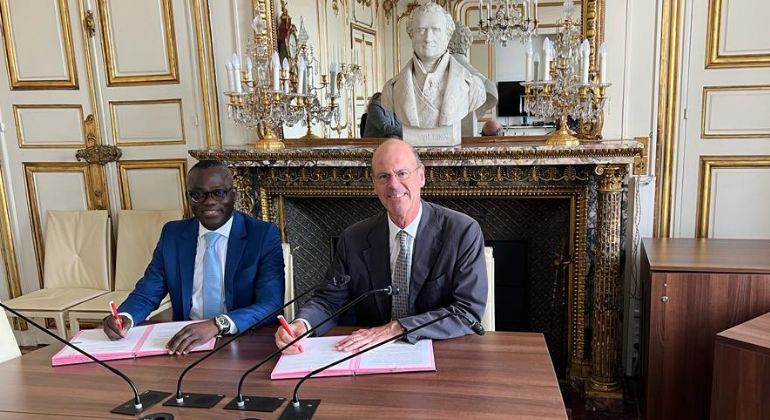 Deux hommes en costume assis à une table signant des documents dans une pièce ornée d'un grand buste et de candélabres dorés